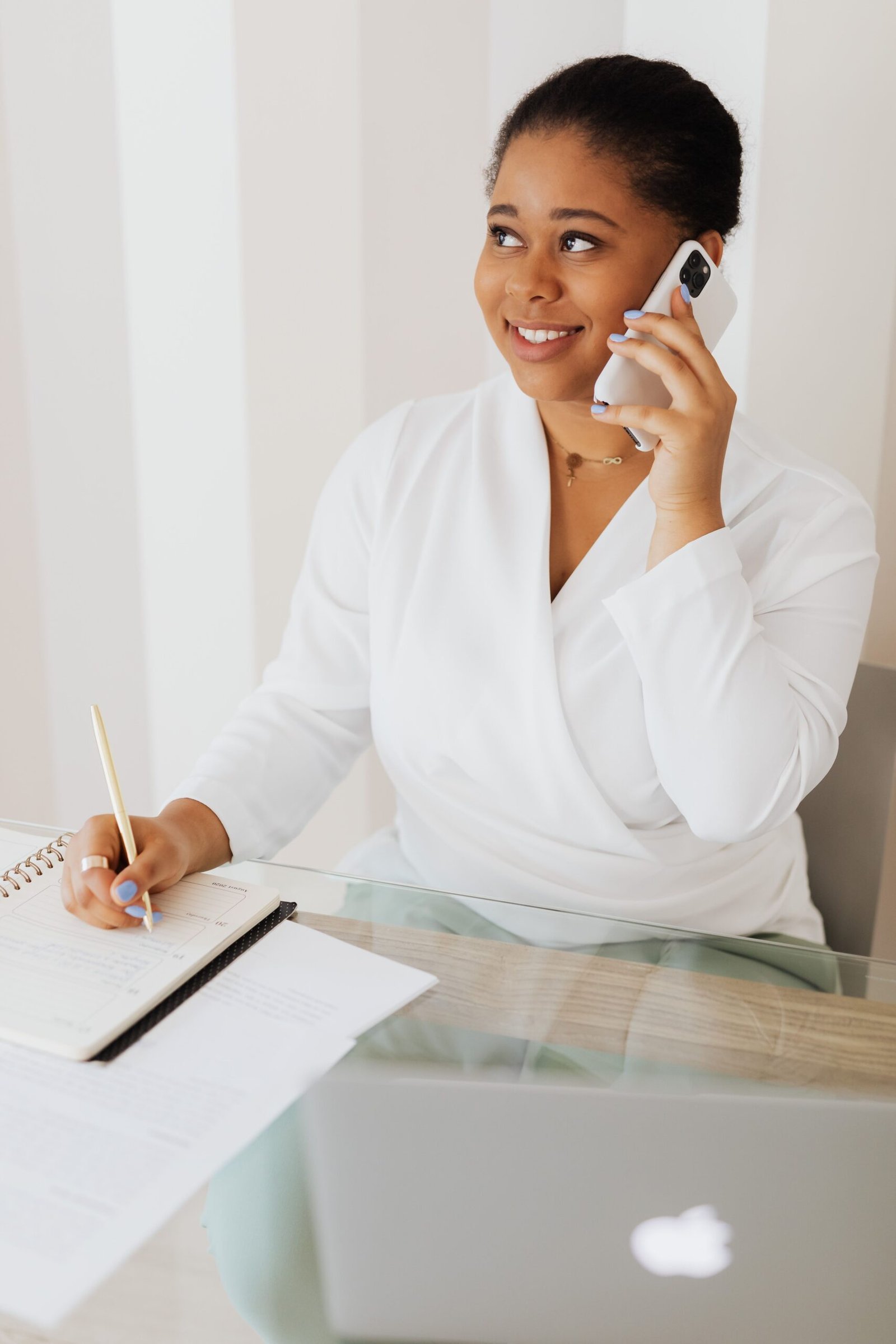 A smiling woman in a white top sits at a desk, using a smartphone and writing notes, conveying a professional setting.